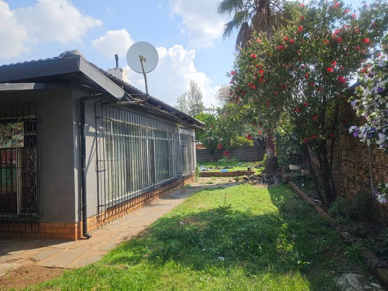 Rear view of the property featuring a paved walkway, security bars on windows, satellite dish, and a garden area with established trees and shrubs.