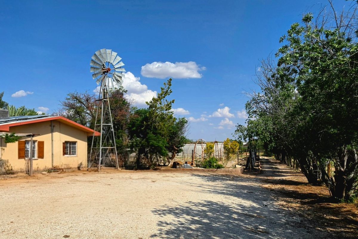 A spacious outdoor area featuring a traditional metal windmill, a gravel driveway, and a greenhouse set within a rural landscape.
