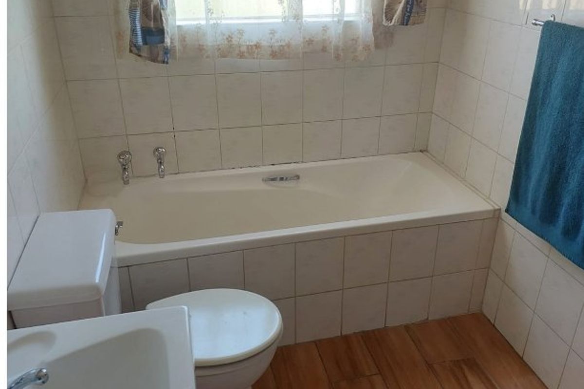 A functional bathroom featuring a built-in bathtub, pedestal washbasin, toilet, and wood-look floor tiles with partial wall tiling.