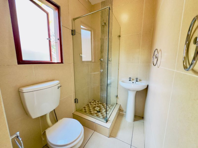 A compact bathroom featuring a pedestal basin, a glass-enclosed shower, and a toilet, finished with neutral wall and floor tiles.