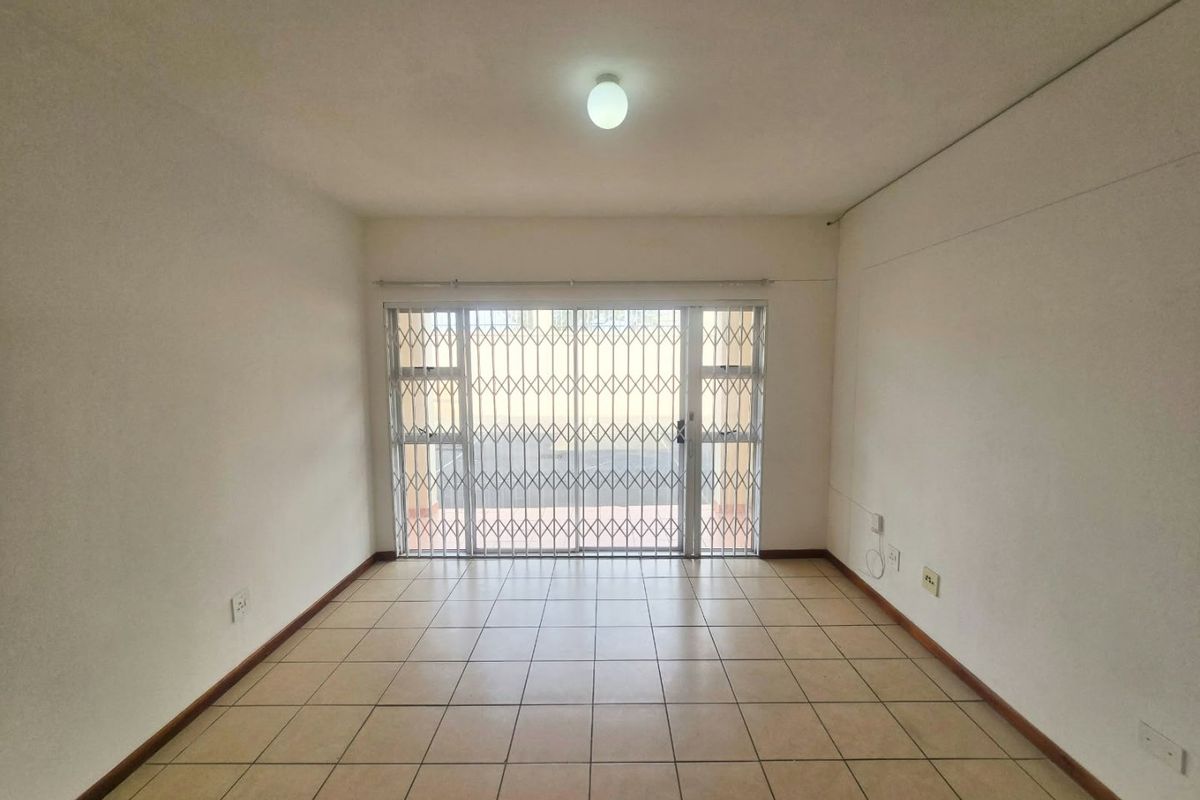 An empty, well-lit living room featuring neutral ceramic floor tiles and a sliding glass door with a security gate leading to the exterior.