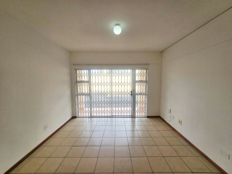An empty, well-lit living room featuring neutral ceramic floor tiles and a sliding glass door with a security gate leading to the exterior.