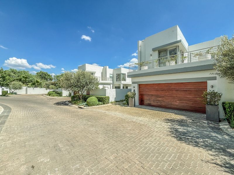 Modern residential exterior featuring a prominent wooden garage door, paved driveway, and a balcony with metal railings under a clear blue sky.