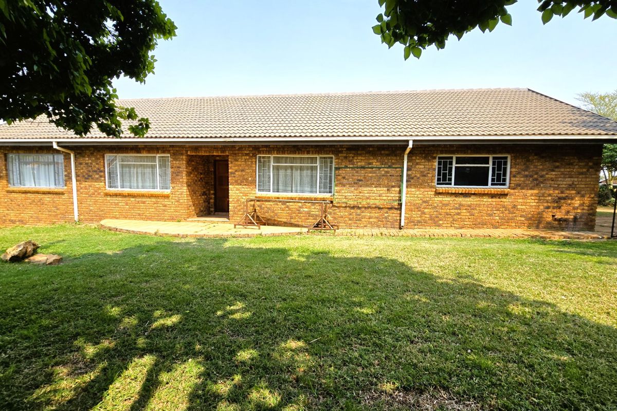 A rear view of a single-story brick house featuring a tiled roof, paved patio area, and a spacious lawn.