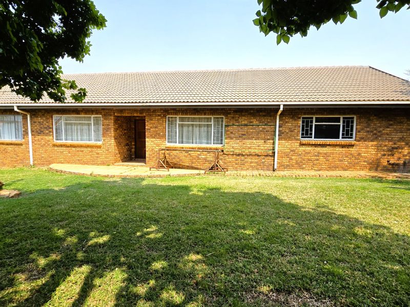 A rear view of a single-story brick house featuring a tiled roof, paved patio area, and a spacious lawn.