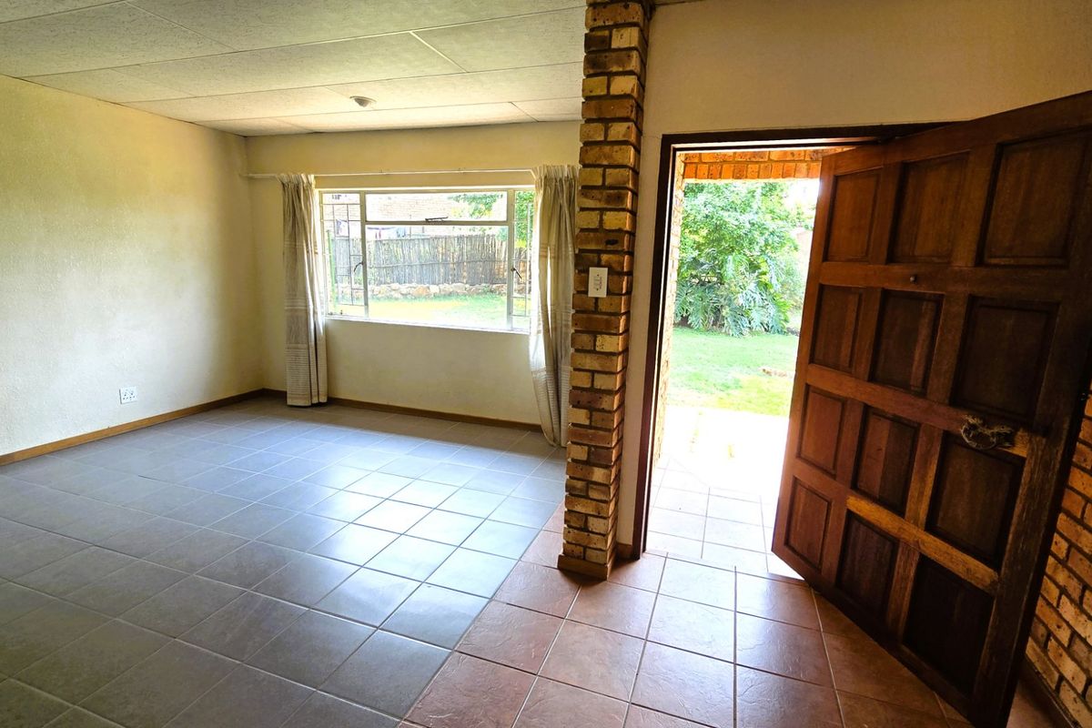 An empty living room featuring tiled flooring, an exposed brick pillar, and direct access to the garden through a solid wood door.
