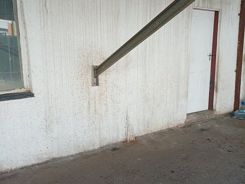 A covered outdoor patio area featuring a concrete floor, steel support structure, and exterior access door, currently showing significant signs of weathering and wear.