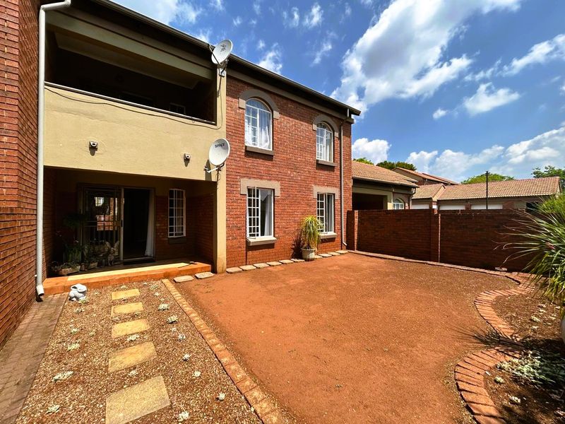 Rear view of a double-story brick apartment featuring a covered patio, arched windows, and a private garden area with paved pathways.