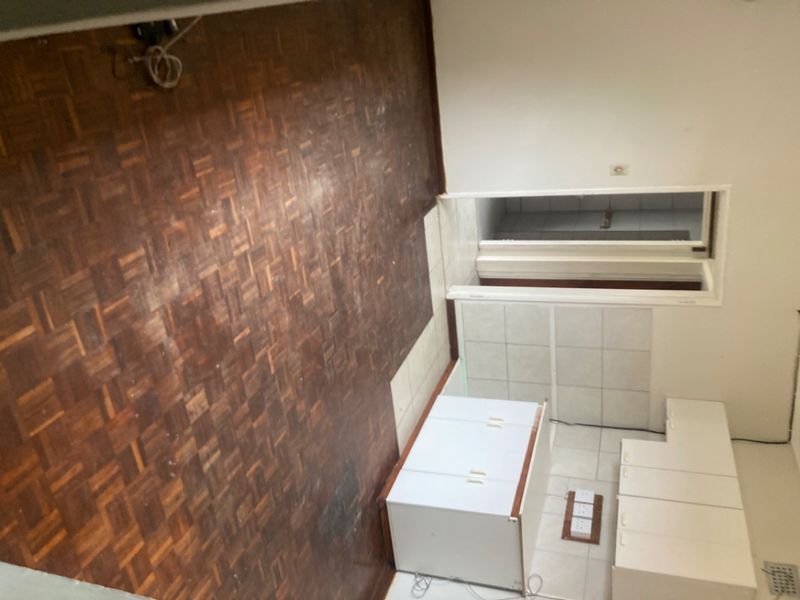 An empty open-plan living and kitchen area featuring original parquet flooring, tiled kitchen zone, and built-in cabinetry, requiring cosmetic updates.
