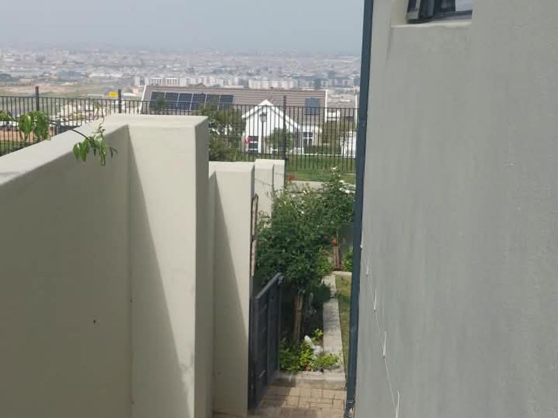 A side pathway featuring paved flooring, boundary walls, and an exterior light fixture, leading towards a rear garden area with distant city views.