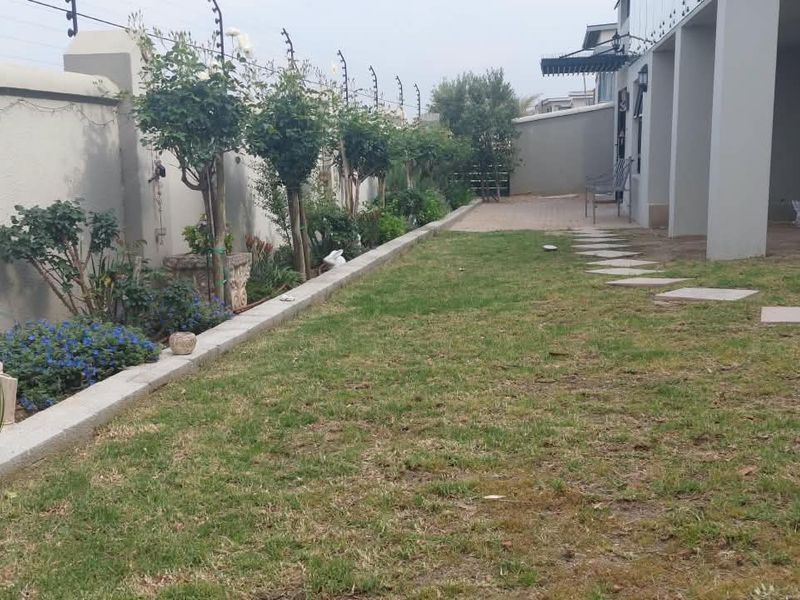 A rear garden area featuring a lawn, paved stepping stones, a covered patio section, and a boundary wall equipped with electric fencing.