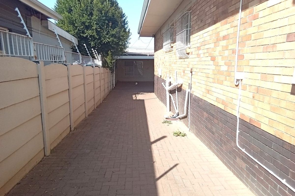 A narrow paved side passage between a brick house wall and a precast concrete boundary wall with security features.