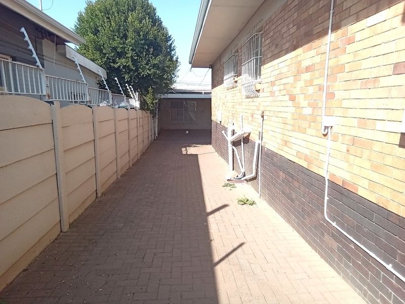 A narrow paved side passage between a brick house wall and a precast concrete boundary wall with security features.
