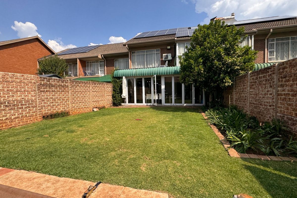 A private rear garden featuring a well-maintained lawn, brick boundary walls, and a paved patio area leading to the townhouse through sliding glass doors.