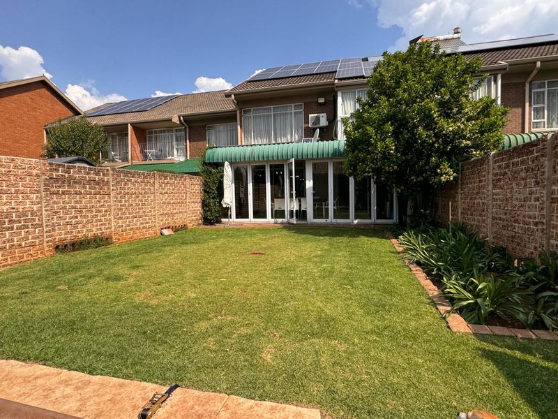 A private rear garden featuring a well-maintained lawn, brick boundary walls, and a paved patio area leading to the townhouse through sliding glass doors.