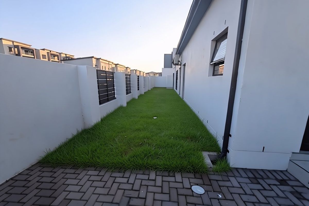 A well-maintained rear garden area featuring a paved walkway, manicured lawn, and secure boundary wall with modern metal fencing inserts.