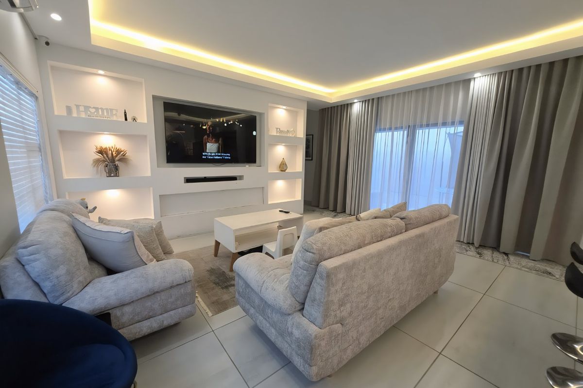 Modern living room featuring a custom built-in media wall with integrated lighting, large format floor tiles, and floor-to-ceiling curtains leading to an outdoor area.