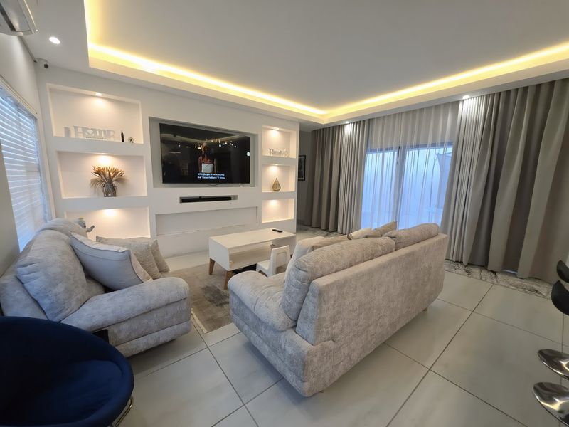 Modern living room featuring a custom built-in media wall with integrated lighting, large format floor tiles, and floor-to-ceiling curtains leading to an outdoor area.