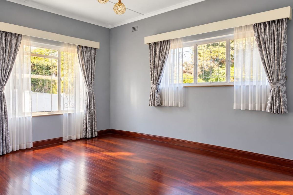 A bright, empty living room featuring polished hardwood flooring, large windows with security bars, and elegant pendant lighting.