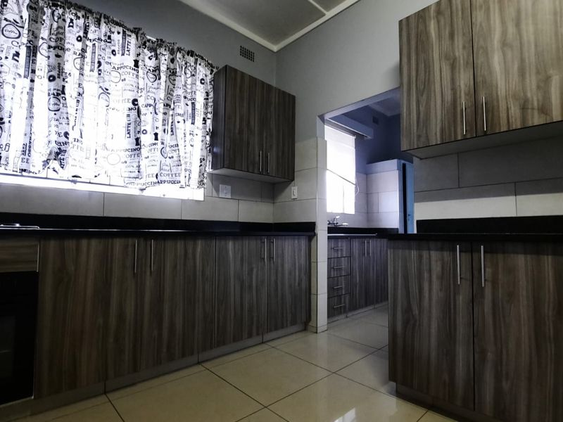 A functional kitchen featuring wood-grain cabinetry, dark countertops, and tiled flooring, with an adjacent scullery area visible through the doorway.
