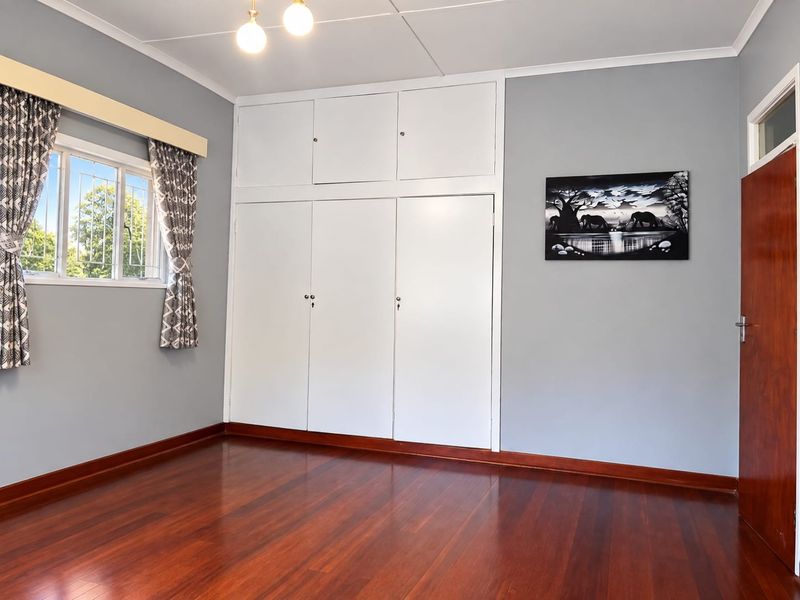 A bright, empty bedroom featuring polished hardwood flooring, built-in floor-to-ceiling cupboards, and security bars on the window.