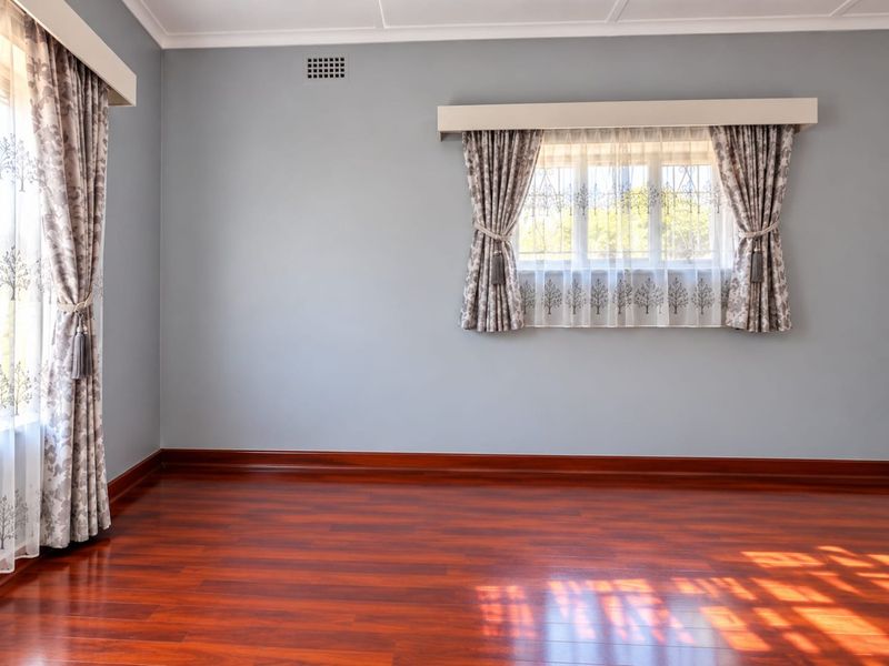 A bright, empty bedroom featuring polished hardwood flooring, neutral grey walls, and windows fitted with security bars and decorative pelmets.