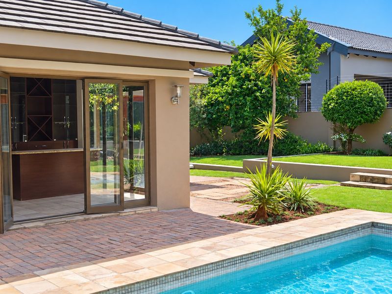 A well-maintained outdoor pool area featuring a paved patio, landscaped garden, and seamless indoor-outdoor flow through large sliding glass doors.