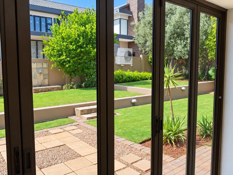 A view through bi-fold doors onto a well-maintained rear garden featuring paved patio areas, tiered lawns, retaining walls, and a decorative wall fountain.