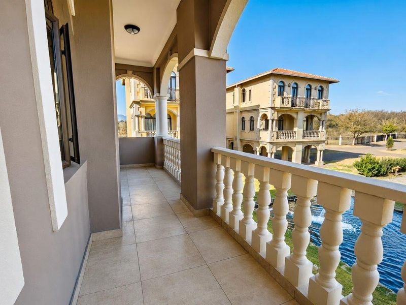 A spacious covered balcony featuring elegant arched columns, a classic balustrade, and neutral tiled flooring, offering pleasant views of the surrounding estate.