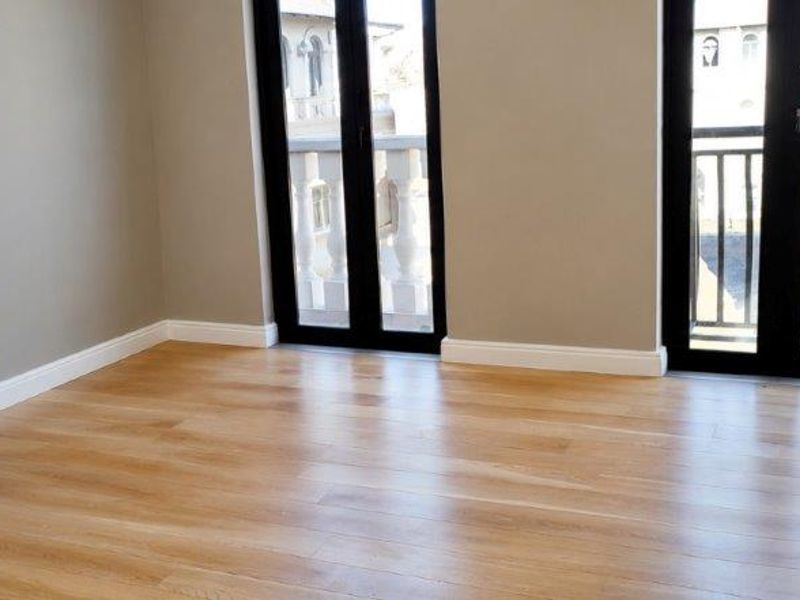 A bright, empty bedroom featuring light wood laminate flooring, neutral walls, and double glass doors leading to a balcony.