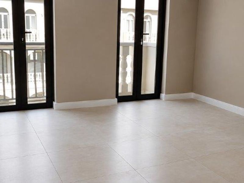A bright, empty bedroom featuring neutral tiled flooring, built-in cupboards, and two sets of double french doors leading to a balcony.