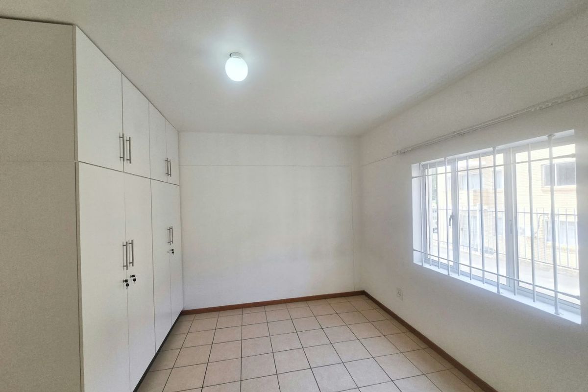 A bright, empty bedroom featuring neutral tiled flooring, built-in white cupboards, and a window fitted with security bars.