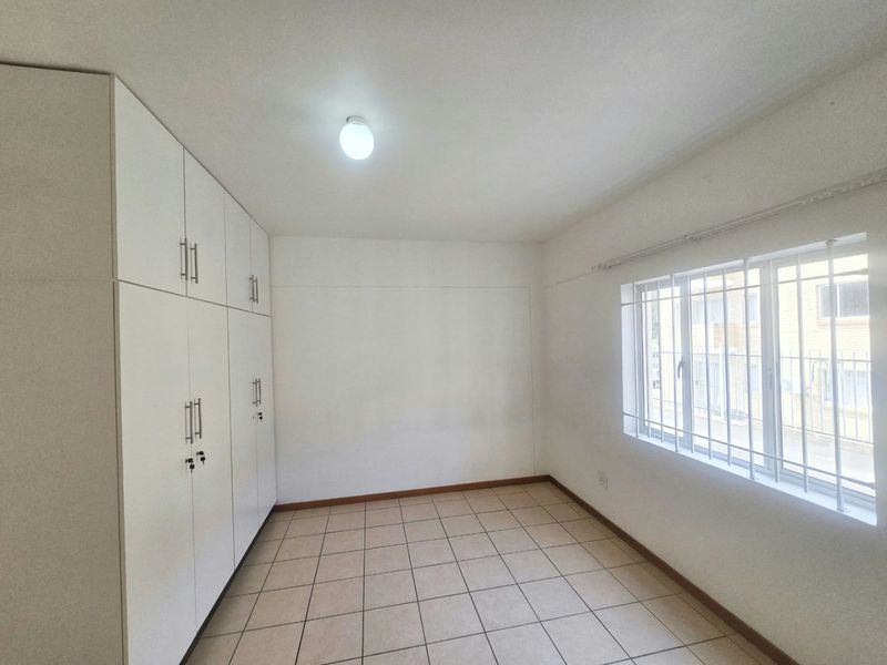 A bright, empty bedroom featuring neutral tiled flooring, built-in white cupboards, and a window fitted with security bars.