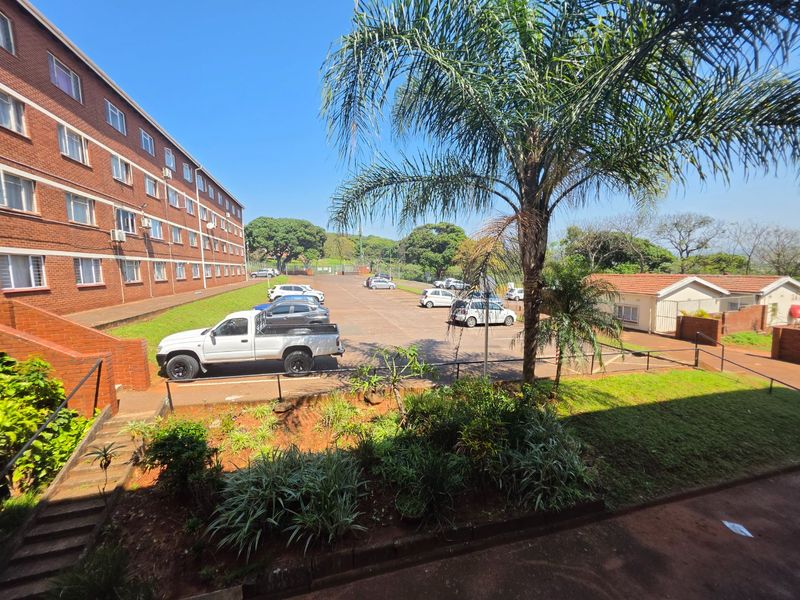 Exterior view of the apartment complex featuring a brick facade, paved parking area, and landscaped garden beds.