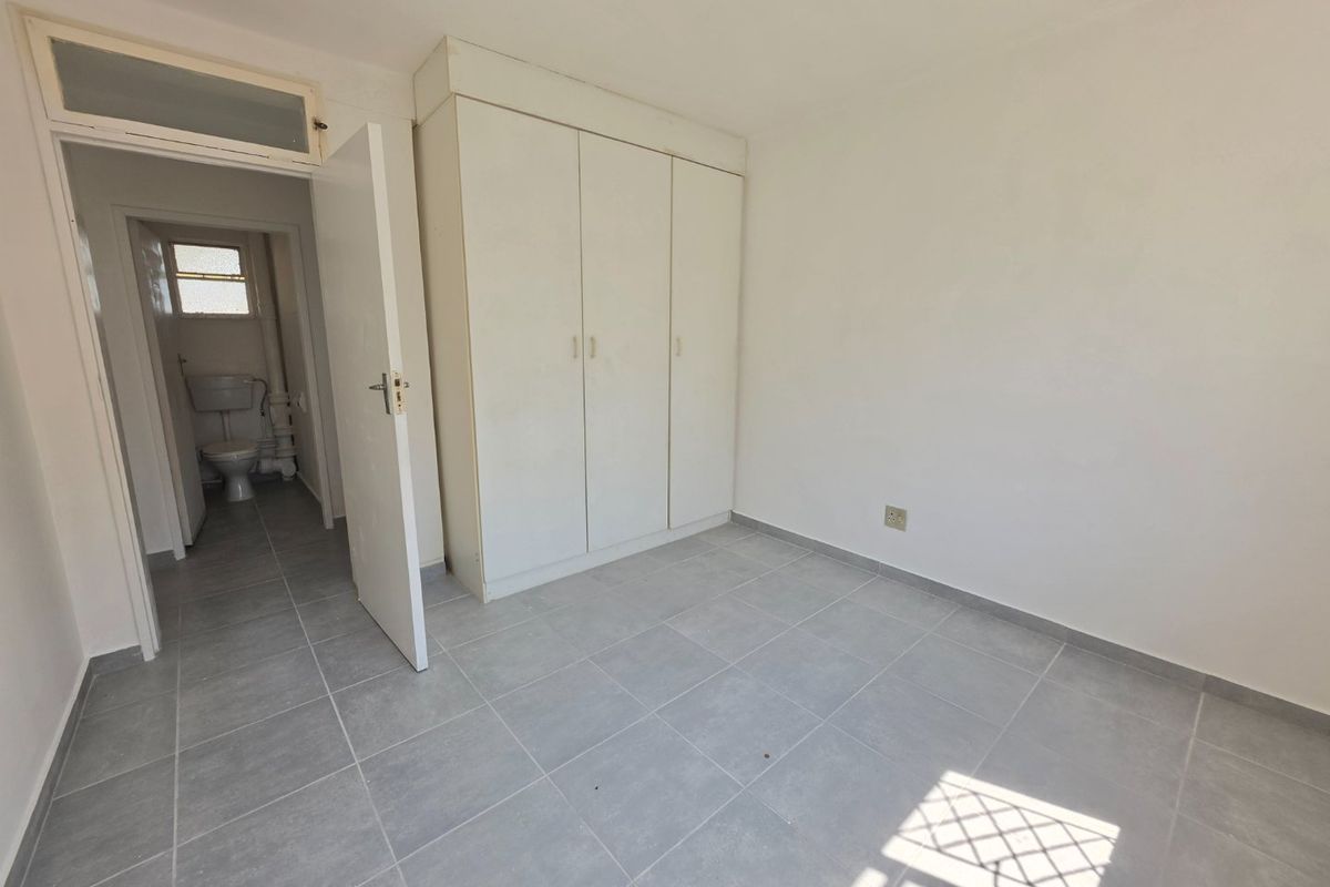 A clean, empty bedroom featuring neutral tiled flooring and a built-in wardrobe, with an adjacent view into a bathroom area.