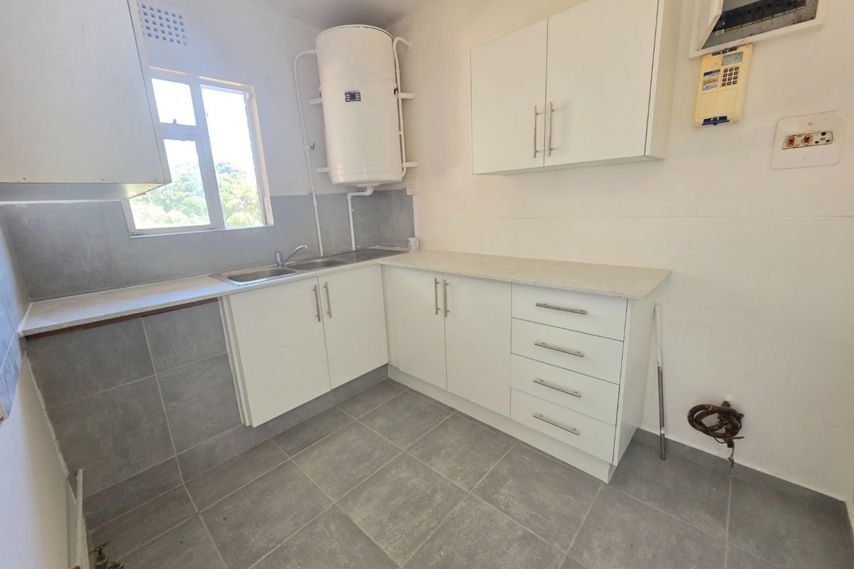 A functional, empty kitchen featuring white cabinetry, grey tiled flooring, a stainless steel sink, and an exposed electric geyser.