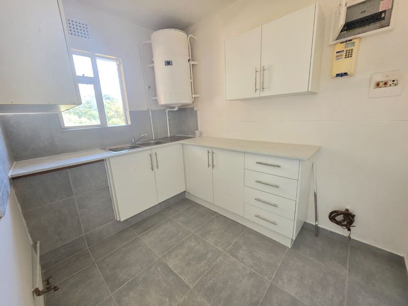 A functional, empty kitchen featuring white cabinetry, grey tiled flooring, a stainless steel sink, and an exposed electric geyser.