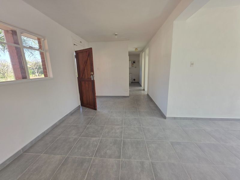 An empty, open-plan living area featuring neutral grey floor tiles, white walls, and a wooden entrance door with natural light from the side window.