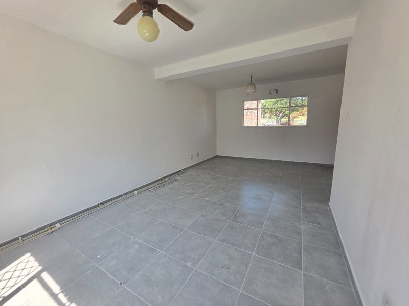 An empty, open-plan living area featuring neutral grey floor tiles, a ceiling fan, and natural light from a large window.