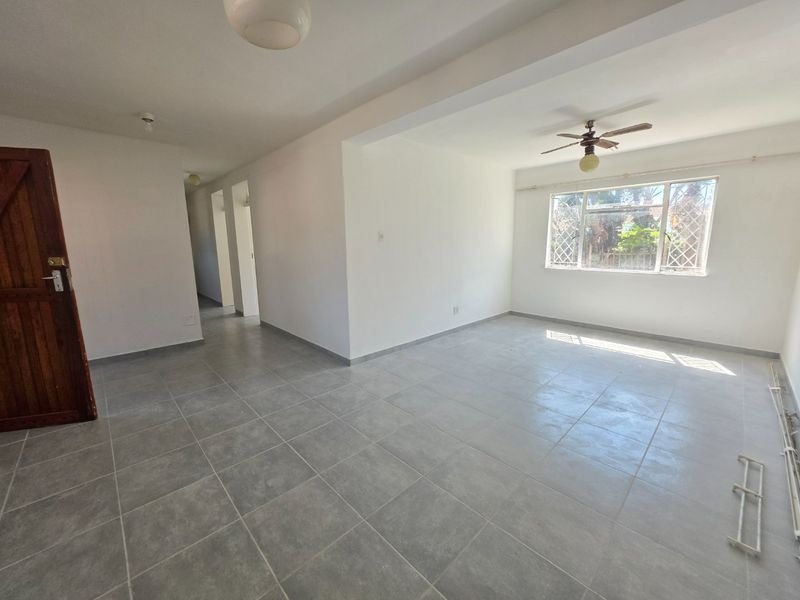 A bright, empty living room featuring neutral tiled flooring, a ceiling fan, and a large window with security bars.