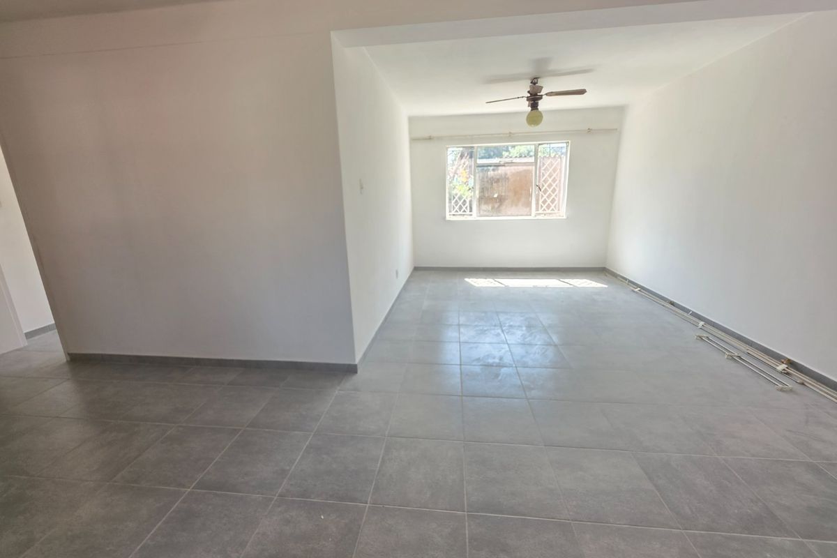 An empty, open-plan living area featuring neutral grey floor tiles, a ceiling fan, and natural light from a large window.