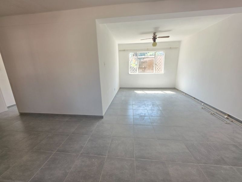 An empty, open-plan living area featuring neutral grey floor tiles, a ceiling fan, and natural light from a large window.