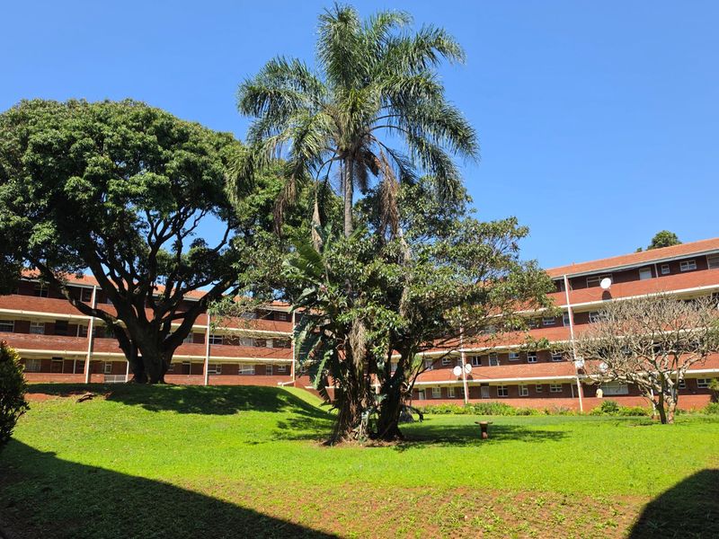 A view of the apartment complex exterior featuring red brick facades, private balconies, and a well-maintained communal garden area with mature trees.
