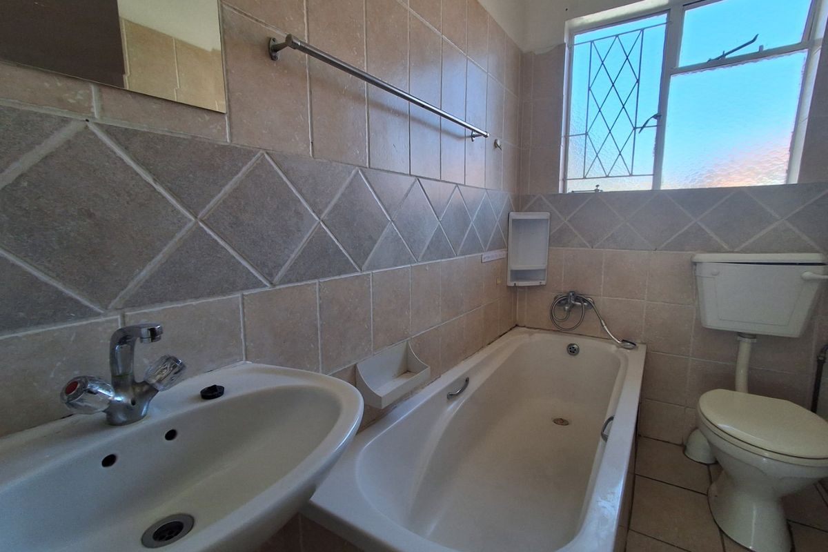 A dated bathroom featuring a standard bathtub, pedestal basin, and toilet with neutral wall tiling and a window with security bars.
