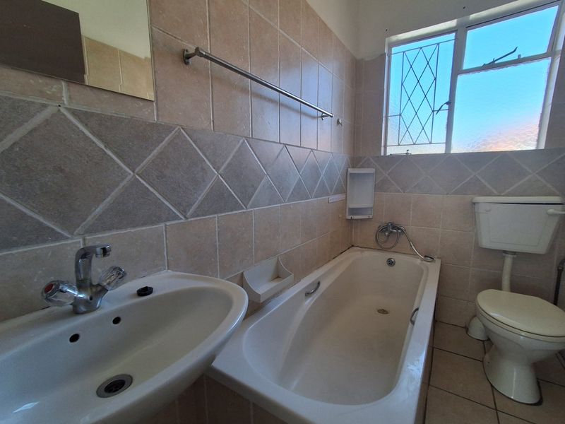 A dated bathroom featuring a standard bathtub, pedestal basin, and toilet with neutral wall tiling and a window with security bars.