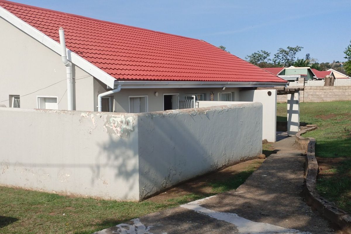 Rear view of a house featuring a red metal roof, a concrete boundary wall with visible wear, and a paved pathway leading to the entrance.