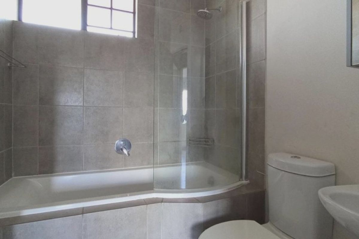 A functional bathroom featuring a bathtub with a glass shower screen, a toilet, and a wall-mounted washbasin with neutral wall tiling.