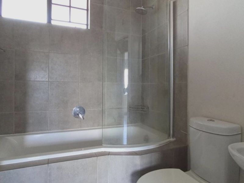 A functional bathroom featuring a bathtub with a glass shower screen, a toilet, and a wall-mounted washbasin with neutral wall tiling.