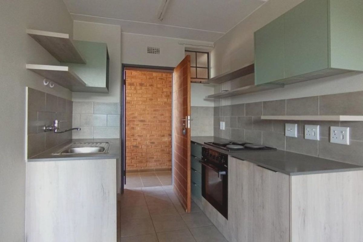 A modern, compact kitchen featuring light wood-finish cabinetry, stone countertops, a built-in oven with hob, and a tiled backsplash.