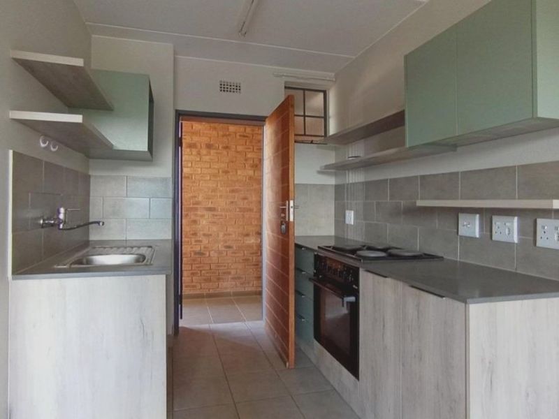 A modern, compact kitchen featuring light wood-finish cabinetry, stone countertops, a built-in oven with hob, and a tiled backsplash.